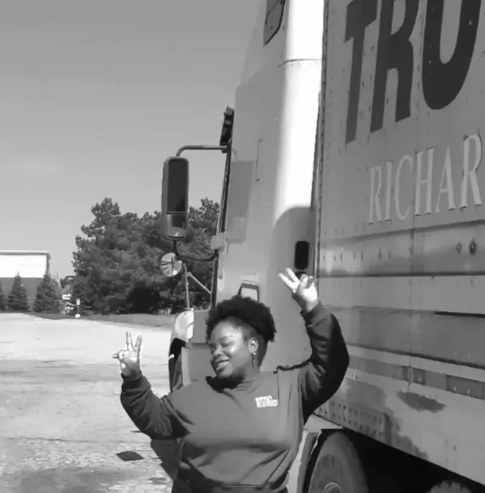 Black and white photo of a woman smiling and giving peace signs beside a semi-truck