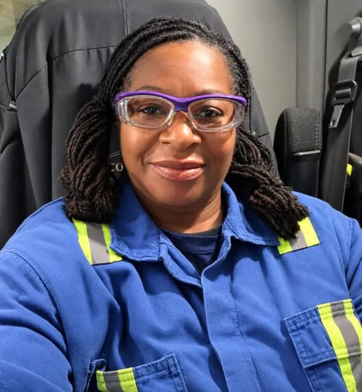 Smiling woman in purple safety glasses and blue hi-vis coveralls sitting in a truck cab