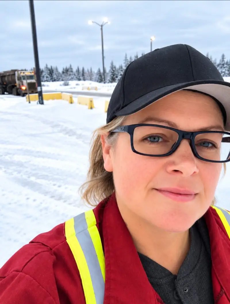 Woman in a cap, glasses and red hi-vis coveralls standing in a snowy truck yard with a truck behind her
